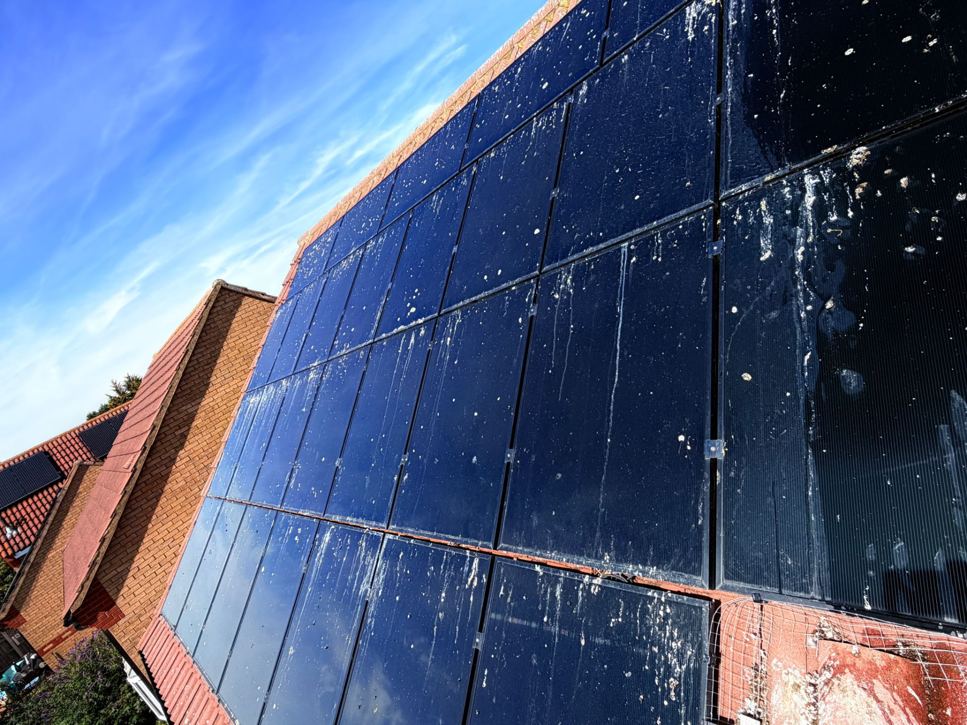 Roof-mounted solar panel array photographed before cleaning, showing white bird droppings, dust and surface grime across the dark panels under a blue sky.