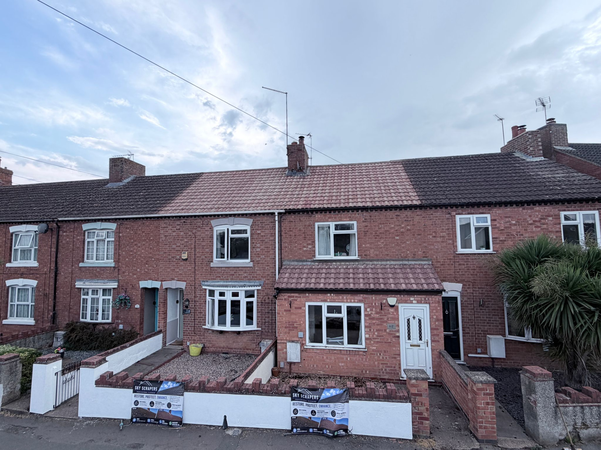 Row of red brick terraced houses photographed from the street, where the middle property has a freshly cleaned red roof while its neighbours remain dark with moss, with Sky Scrapers Ltd boards at the garden wall.